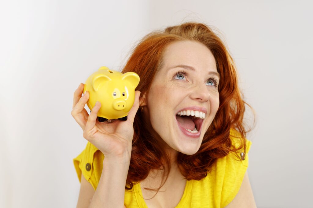 Eine fröhliche, rothaarige Frau in einem gelben T-Shirt hält ein gelbes Sparschwein und lacht begeistert. Das Bild symbolisiert finanzielle Unabhängigkeit und Freude über erfolgreiches Sparen