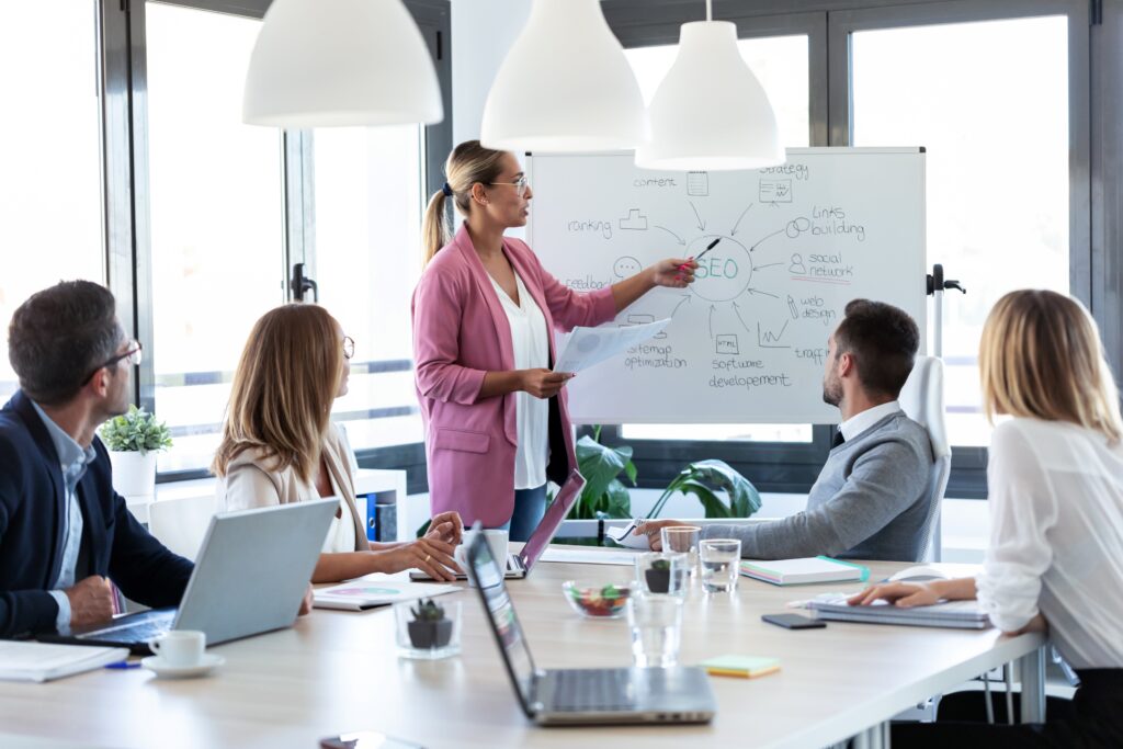 Eine Frau in einem rosa Blazer leitet eine SEO-Schulung für ein Team in einem modernen Büro. Sie erklärt verschiedene SEO-Strategien auf einem Whiteboard, während die Teammitglieder aufmerksam zuhören und Notizen machen. Das Bild zeigt die Bedeutung von SEO-Kursen zur Verbesserung der Online-Sichtbarkeit und des Geschäfts