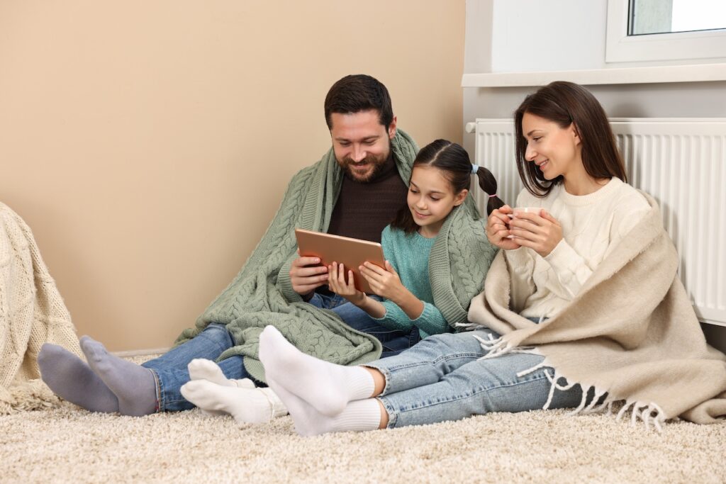 Eine Familie, eingehüllt in Decken, sitzt gemütlich vor der Heizung auf dem Teppich und schaut zusammen auf ein Tablet.