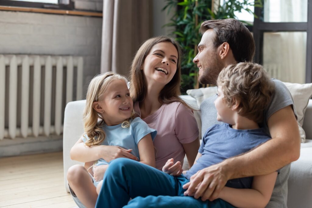 Eine lachende Familie sitzt gemeinsam im Wohnzimmer, mit einem Heizkörper im Hintergrund und warmem Tageslicht.