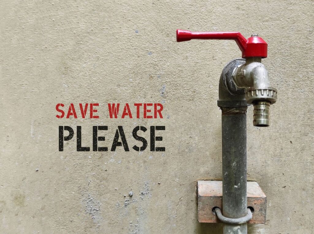 Wasserhahn mit rotem Griff vor Betonwand mit Aufschrift save water please, Symbol fuer Wassersparen und nachhaltige Nutzung mit zisterne